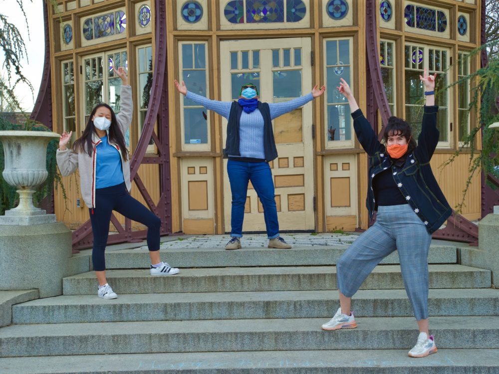 Image of 3 people in masks outside on steps with their arms in the air